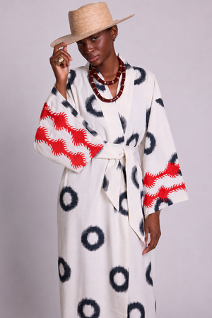 A model wearing a KLEED white and black kimono with red motifs embroideres on the sleeves, styled with a straw hat and layered red beaded necklaces against a light gray background