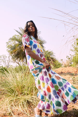 Editorial photo of a model standing in tall grass beneath palm trees, wearing the Anouk Bikaner Dress by Kleed, a tiered maxi dress in 100% handloom cotton ikat with a multicoloured pattern on a white base, with short sleeves and a flounced hem.