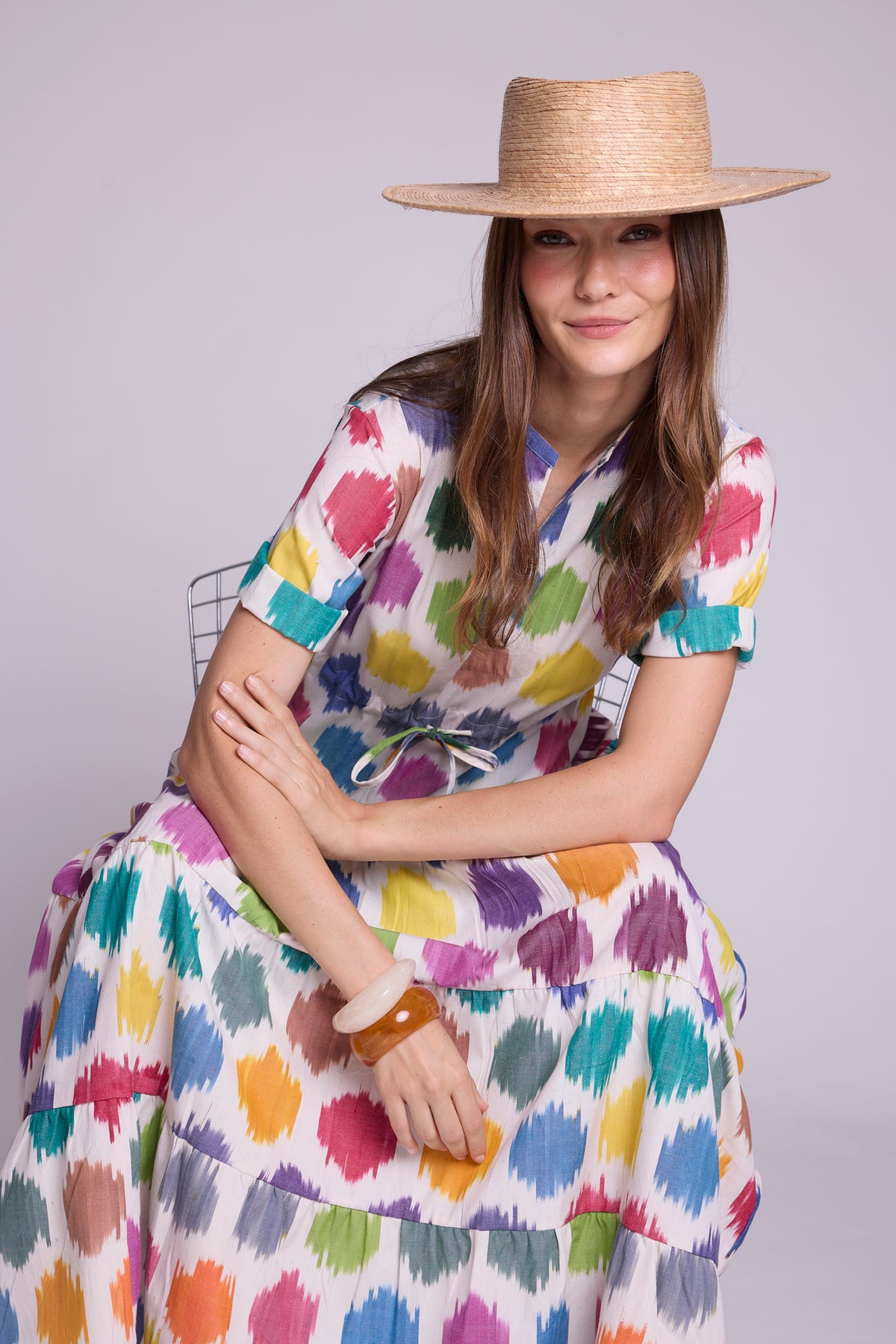 Model seated wearing the anouk maxi dress by kleed in 100% cotton ikat, colourful geometric motifs and some bracelets and straw hat.