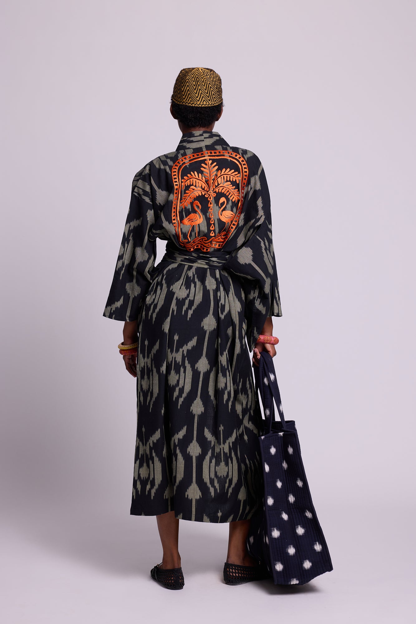 Model wearing a long cotton ikat kimono by kleed  with black and khaki geometric pattern with 2 orange flamingos and a palm tree embroidered at the back.