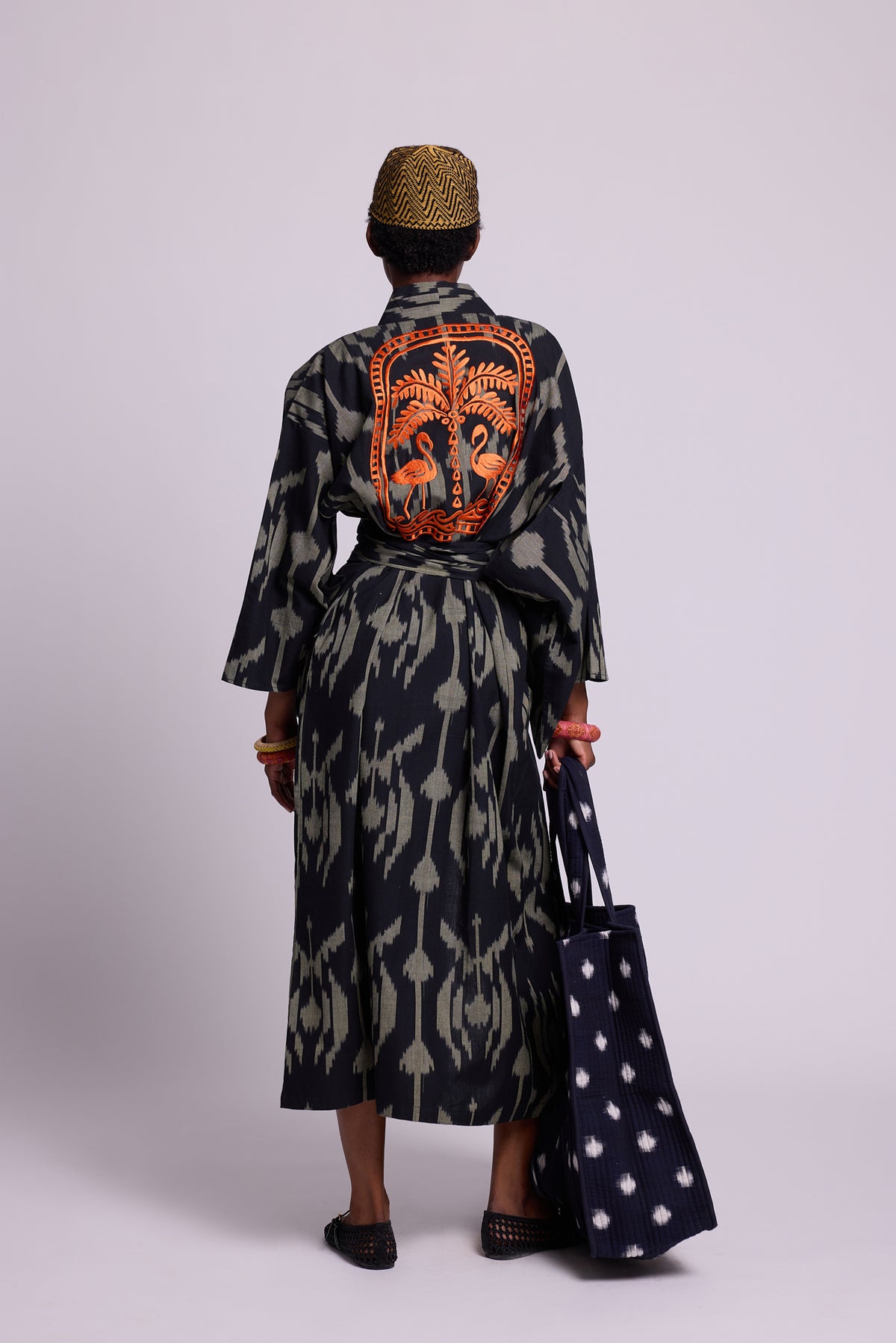 Model wearing a long cotton ikat kimono by kleed  with black and khaki geometric pattern with 2 orange flamingos and a palm tree embroidered at the back.