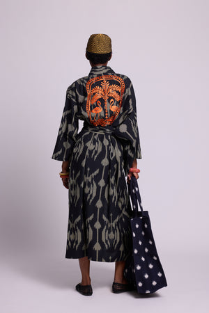Model wearing a long cotton ikat kimono by kleed  with black and khaki geometric pattern with 2 orange flamingos and a palm tree embroidered at the back.