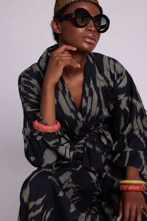 Woman wearing a cotton ikat kimono by kleed with black and khaki geometric pattern black sunglasses and colourful bracelets against a plain background