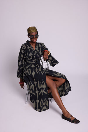 Model wearing a patterned cotton ikat kimono in black and khaki geometric patterns sitting on a chair against a white background with black sunglasses and colourful bangles