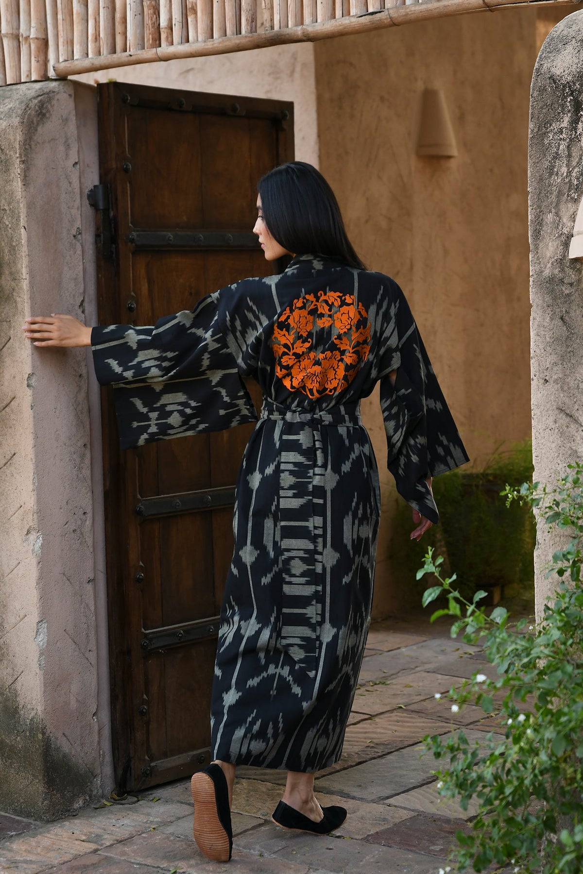 Woman wearing Kleed Congo Naya Kimono in handloomed Ikat cotton black and khaki abstract pattern opening wooden door