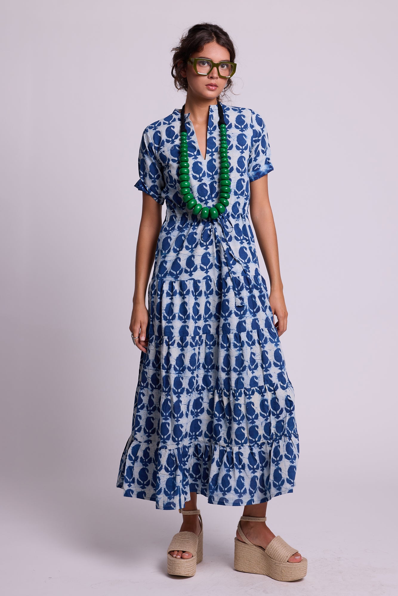 A woman modeling a blue and white patterned short-sleeve maxi dress the crete bikaner by Kleed, hand blockprinted in 100% chemical free cotton. With a green beaded necklace and beige platform straw sandals, standing against a light gray studio background.