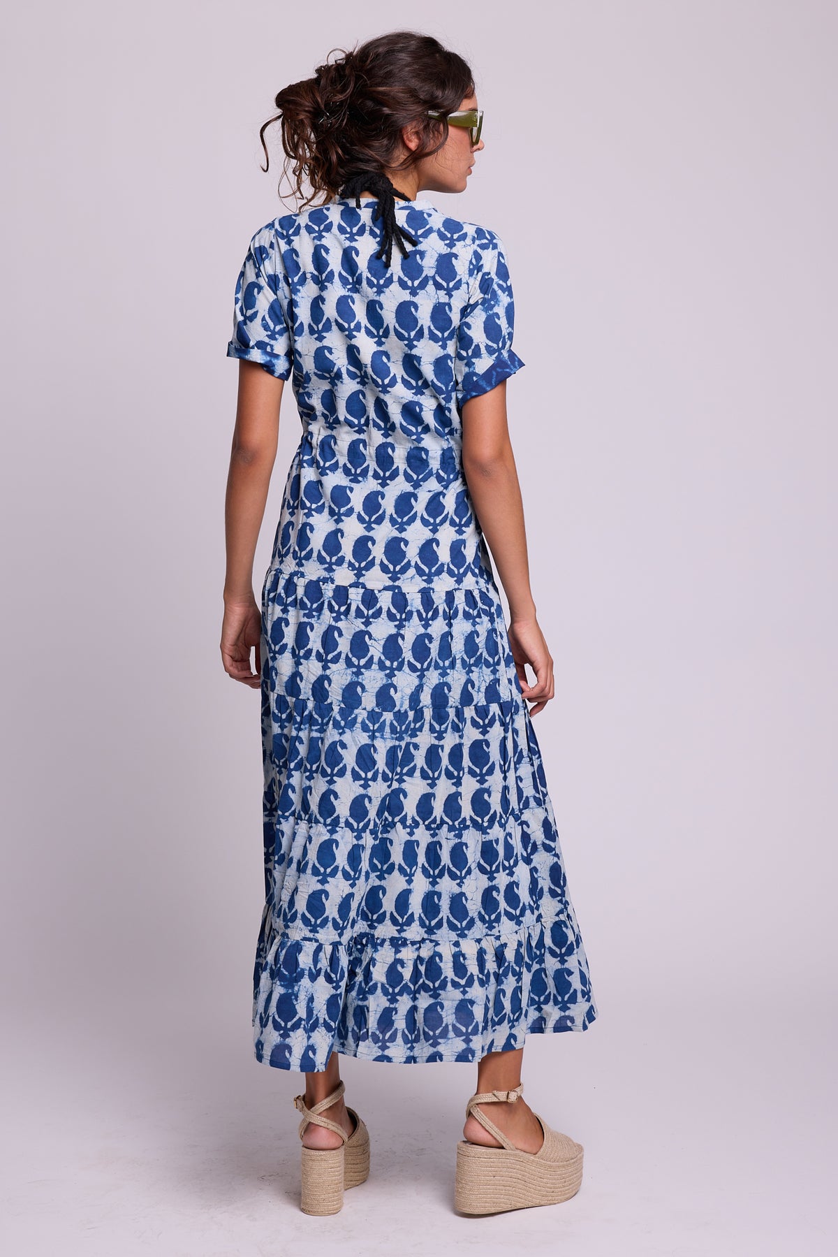 A woman wearing a short-sleeve blue and white patterned maxi dress, the crete bikaner by Kleed, hand blockprinted in 100% chemical free cotton, shown from the back against a light gray studio background.