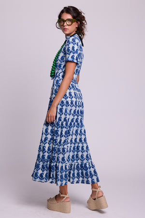 A woman modeling a short-sleeve maxi dress with a indigo blue and white hand block printed in 100% cotton,  styled with a green beaded necklace and beige platform sandals against a solid light gray studio background.