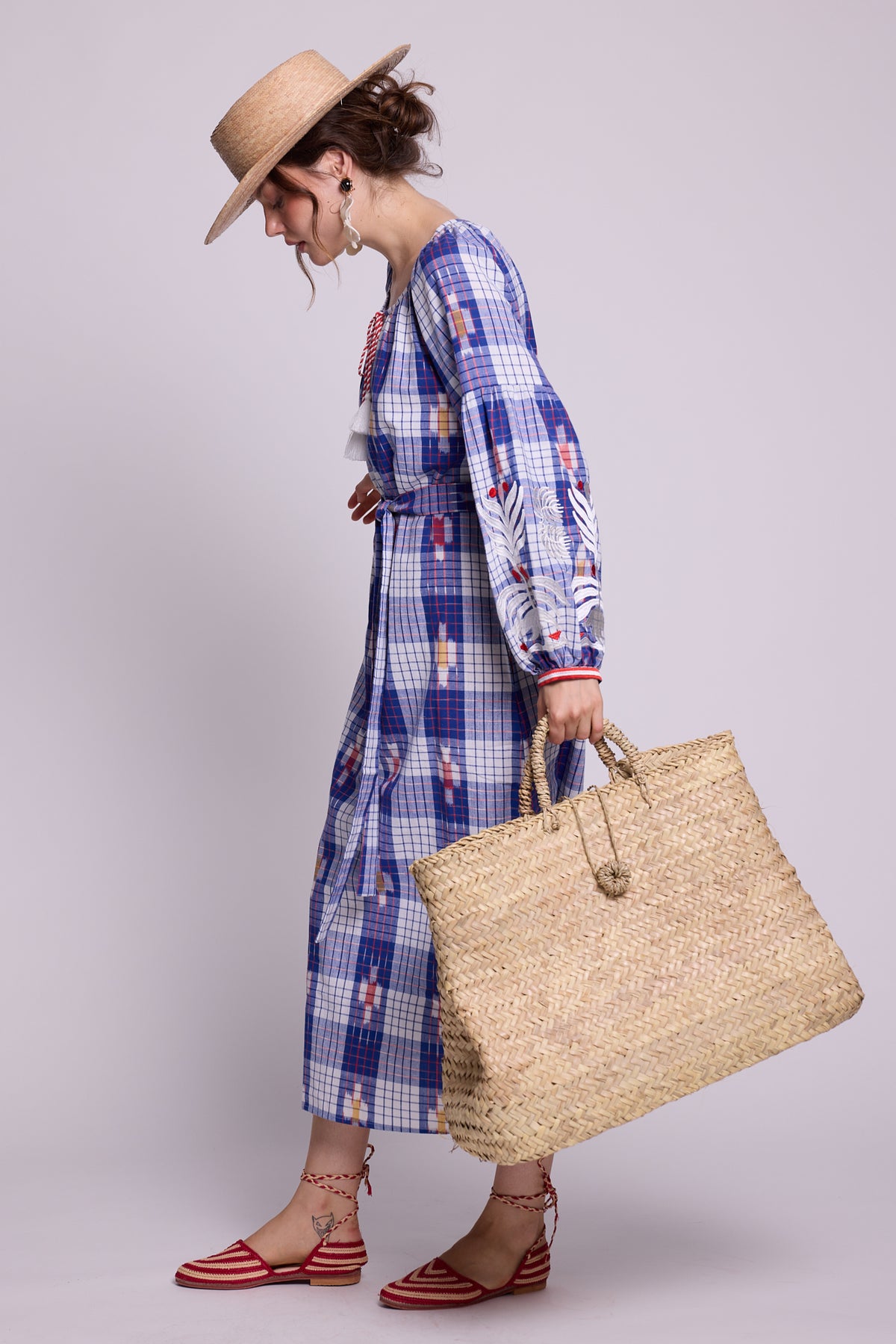 A woman in a straw hat and red-and-white striped sandals models a blue and white plaid cotton kaftan by kleed, produced in hand looms in ikat pattern, holding a large woven straw tote bag against a solid light gray background.