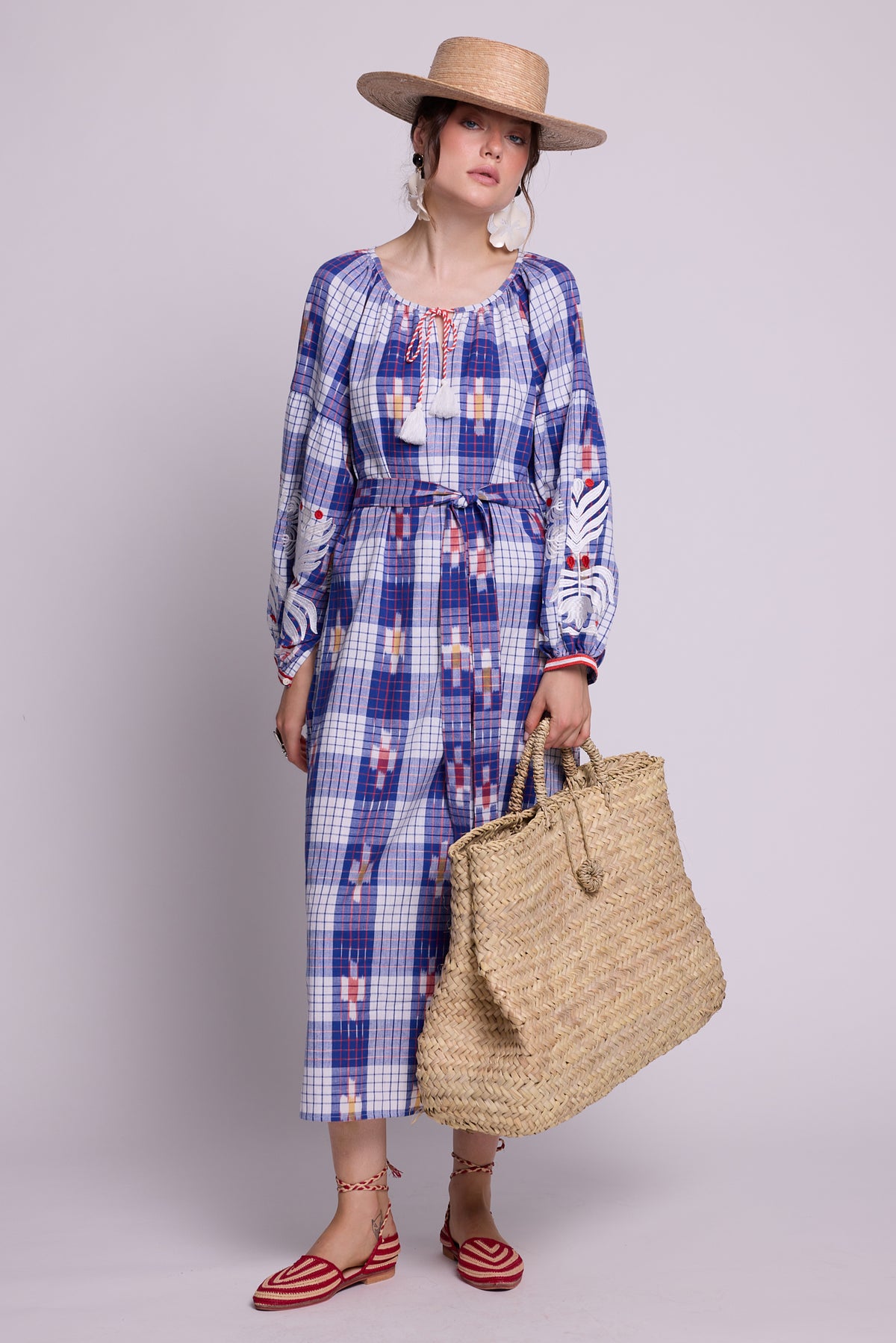 A model wearing the french kaftan by kleed, produced in hand looms, in 100% ikat, with checks blue red and white, embroidered in white on the sleeves and red and white on the cuffs.  A straw hat, holding a straw basket and white floer earings.