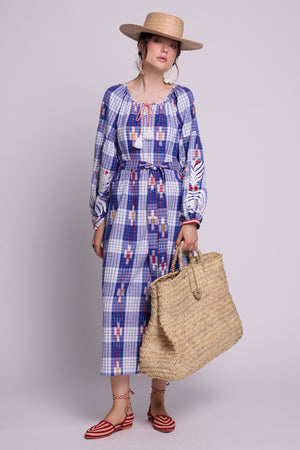A model wearing the french kaftan by kleed, produced in hand looms, in 100% ikat, with checks blue red and white, embroidered in white on the sleeves and red and white on the cuffs.  A straw hat, holding a straw basket and white floer earings.