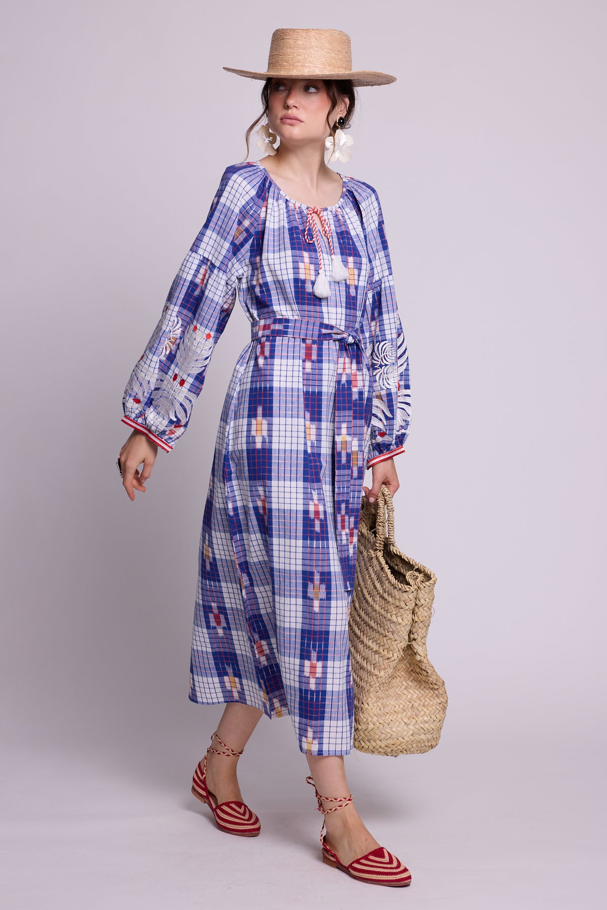 A woman modeling a long-sleeve hand woven ikat kaftan by kleed, with a blue, white, and red check pattern, styled with a straw hat, a woven straw bag, and red-and-white striped sandals against a light gray studio background. A slow loungewear iece for summer.