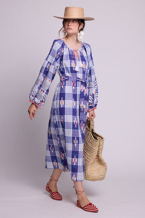 A woman modeling a long-sleeve hand woven ikat kaftan by kleed, with a blue, white, and red check pattern, styled with a straw hat, a woven straw bag, and red-and-white striped sandals against a light gray studio background. A slow loungewear iece for summer.
