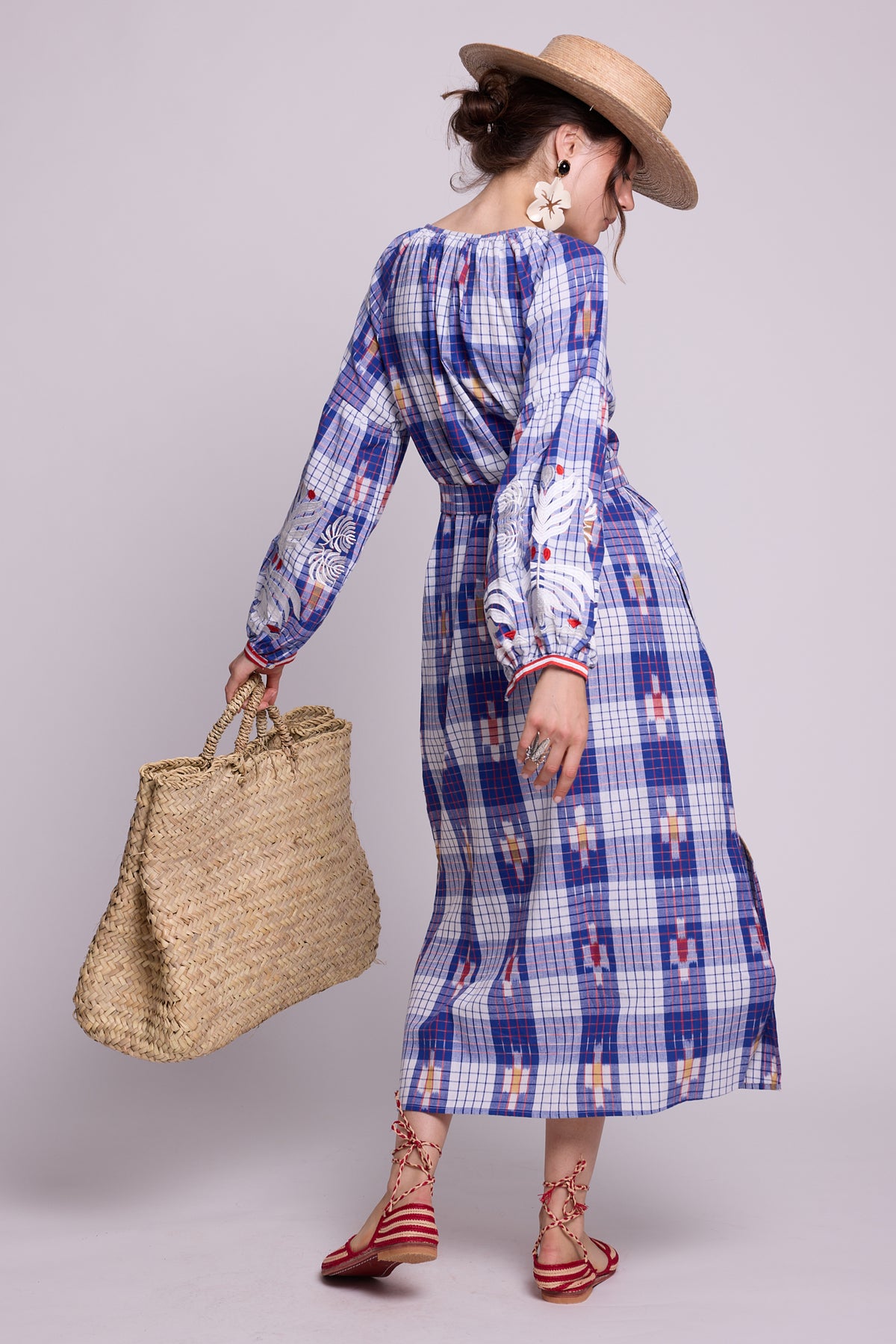 A woman wearing a blue and white gingham ikat cotton kaftan, yhe french kaftan by kleed, with white embroidered details on the sleeves and red and white embroidred cuffs, holding a large woven straw tote bag. She is wearing a straw hat and red sandals, photographed against a solid light gray studio background.