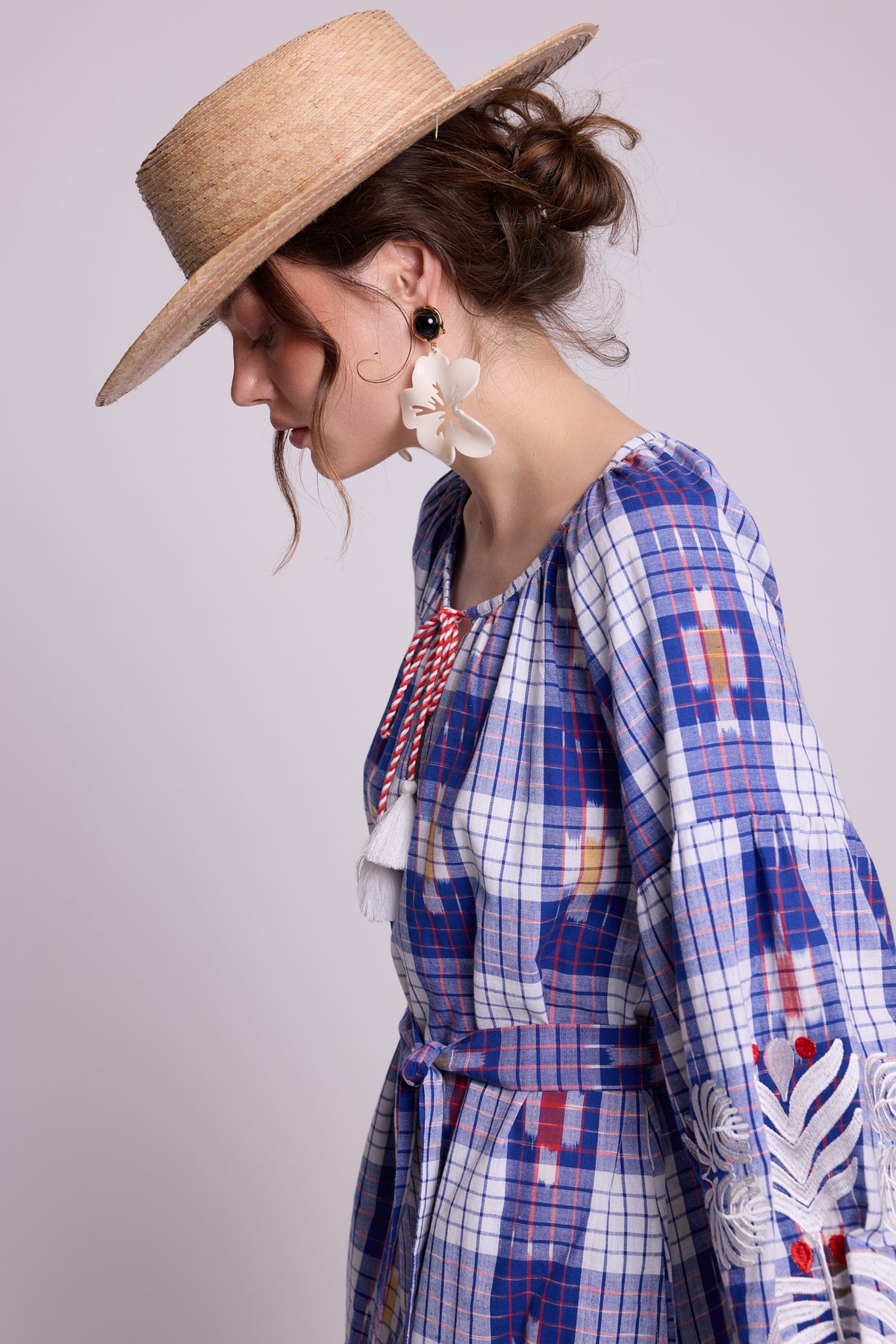 A woman in profile wearing a straw hat, white flower earrings, and a blue, white, and red plaid kaftan by kleed the frenck kaftan, produced with 100% cotton ikat, with embroidered sleeves, set against a solid light gray background.