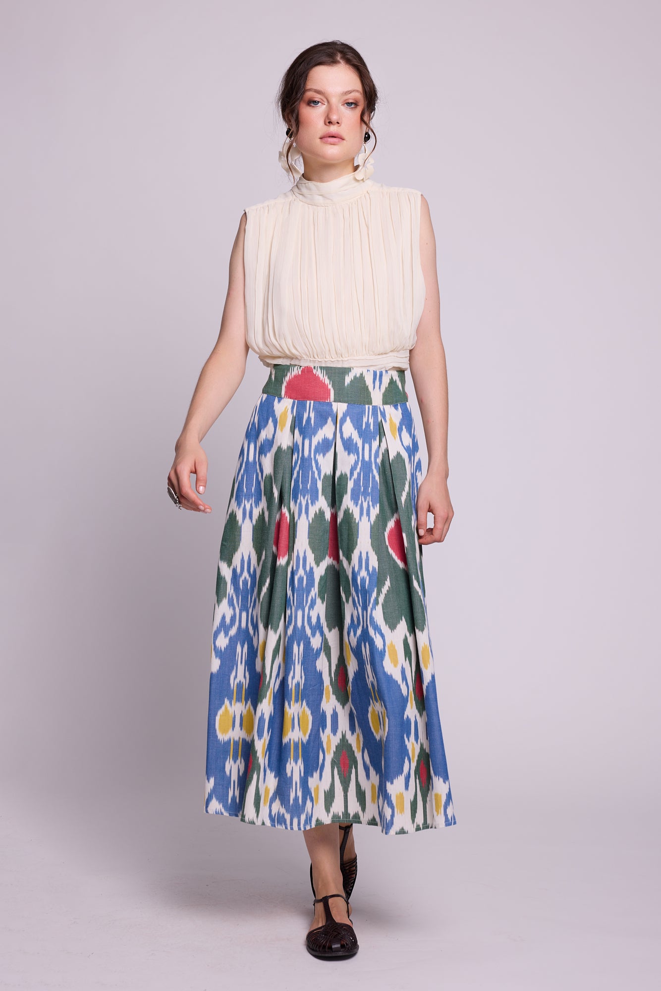 A woman modeling a sleeveless cream pleated top paired with the garden skirt by kleed a high-waisted midi skirt featuring a blue, green, red, and white ikat pattern, set against a solid light gray studio background.