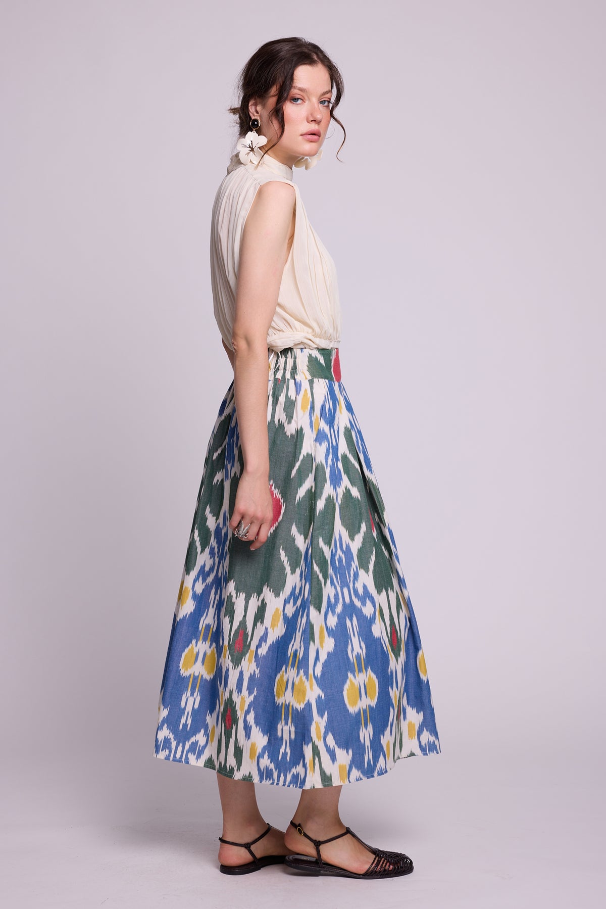 A woman modeling a sleeveless cream top and  teh garden skirt by kleed, a high-waisted midi skirt with a blue, green, and yellow ikat pattern, standing against a light gray studio background.