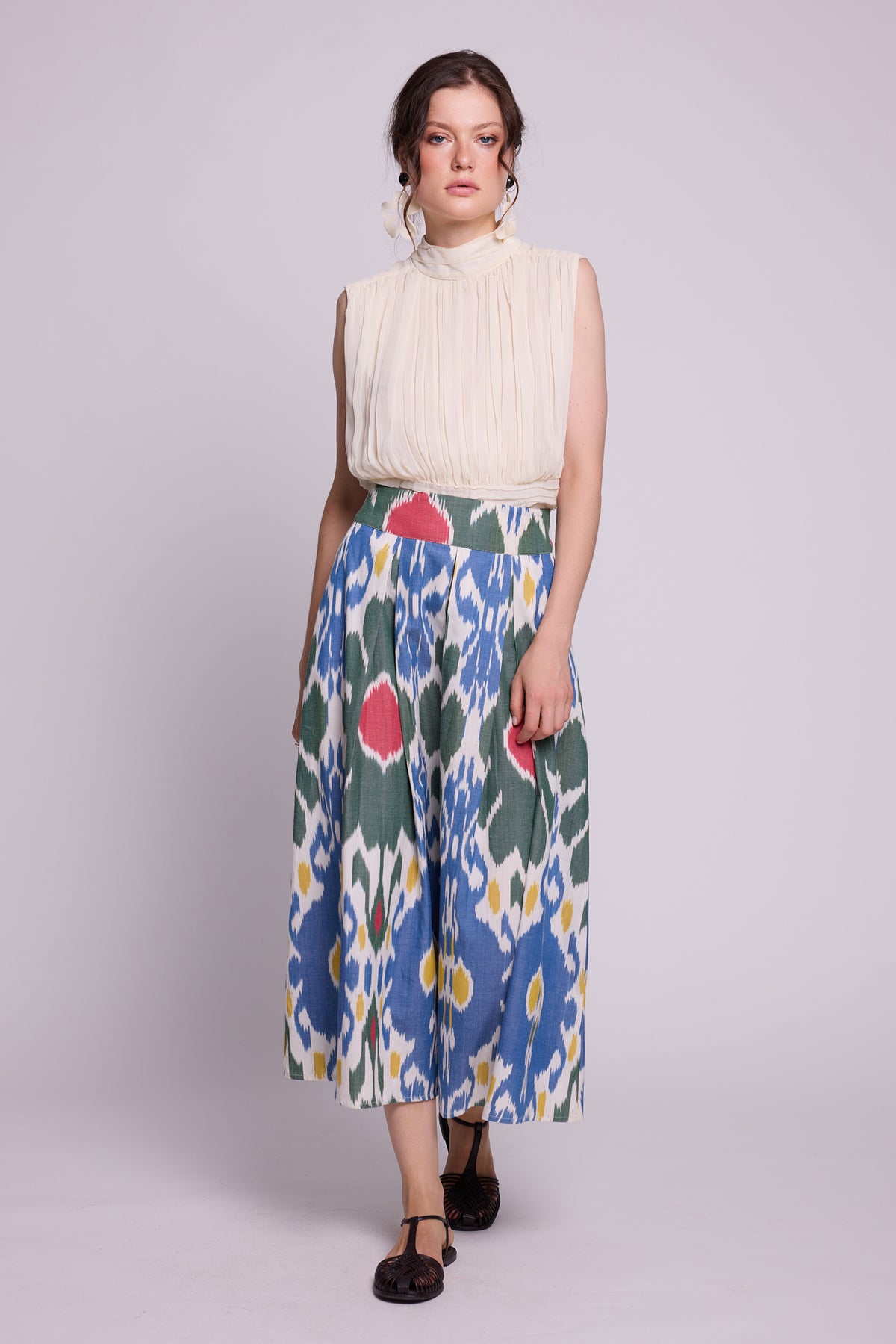 A model wearing a sleeveless cream high-neck top and the garden skirt by kleed,  with a blue, green, red, and yellow ikat pattern, standing against a light gray studio background.