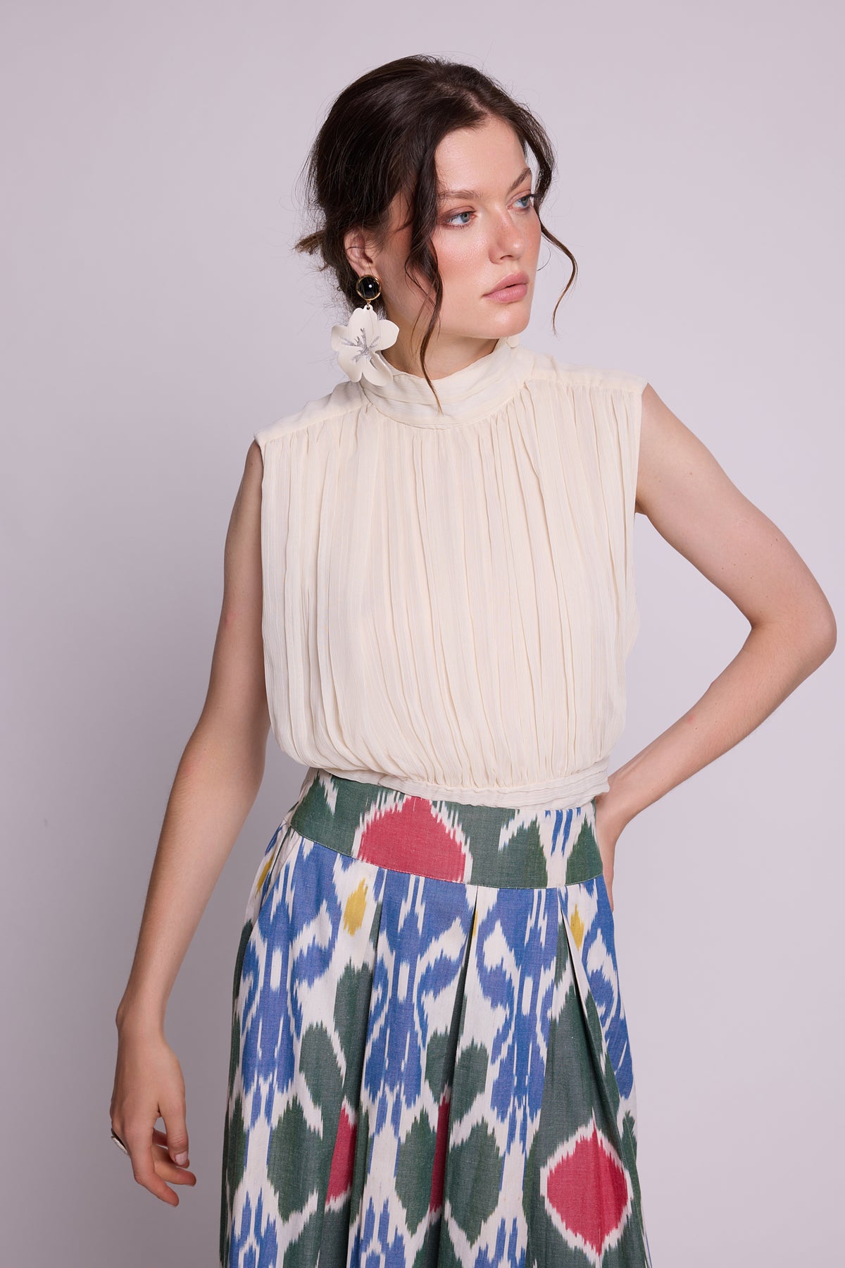 close up of a woman modeling a sleeveless cream pleated top paired with athe garden skirt by kleed, a colorful patterned skirt featuring blue, green, red, and yellow ikat-style motifs against a light gray studio background.