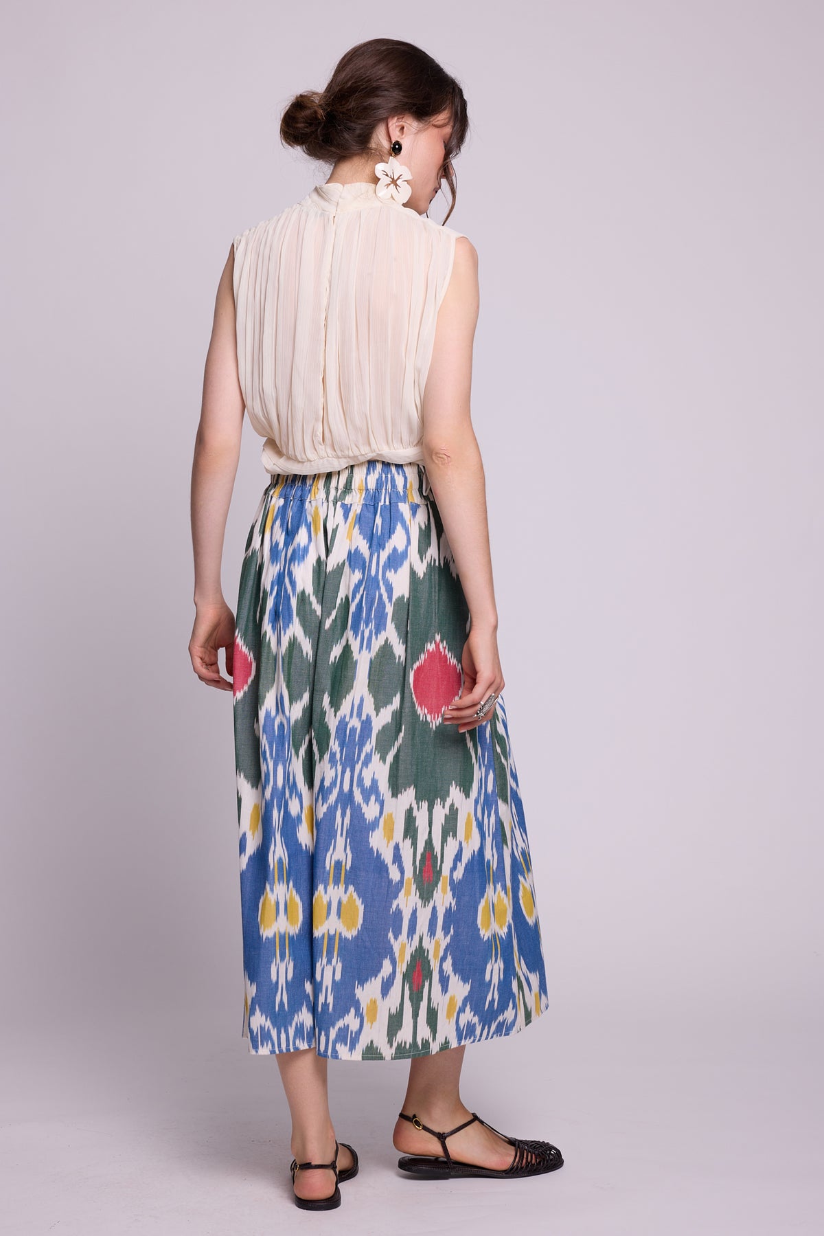 A woman shown from the back wearing a sleeveless cream pleated top and the garden skirt by kleed a high-waisted midi skirt with a blue, green, and white ikat floral pattern. She is standing against a light gray studio background. 