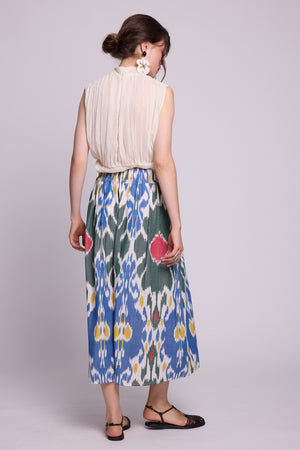 A woman shown from the back wearing a sleeveless cream pleated top and the garden skirt by kleed a high-waisted midi skirt with a blue, green, and white ikat floral pattern. She is standing against a light gray studio background. 