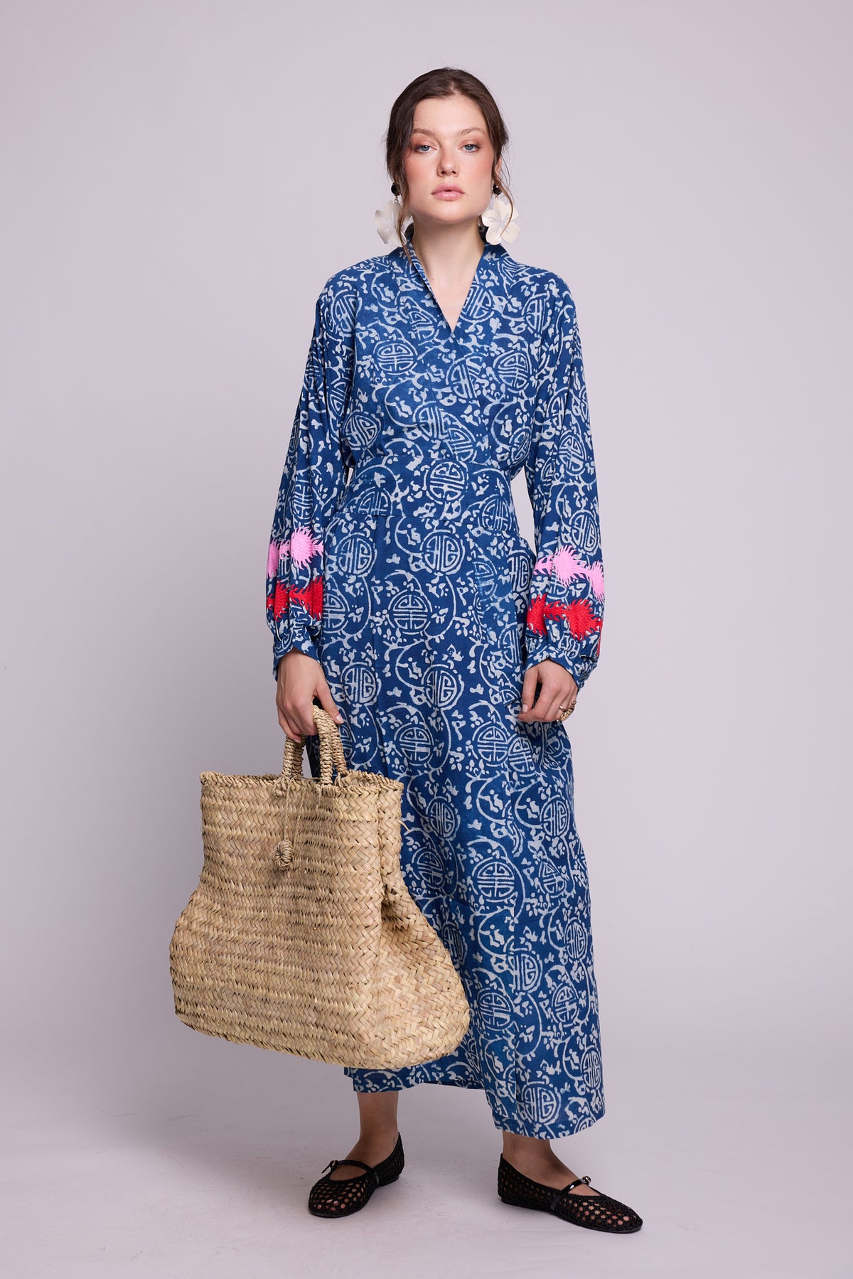 A woman modeling a long balloon sleeved indigo blue and white patterned handblock printed kimono by Kleed, 100% chemical free, with red and pink embroidered details on the cuffs, holding a large woven straw tote bag against a solid light gray background.
