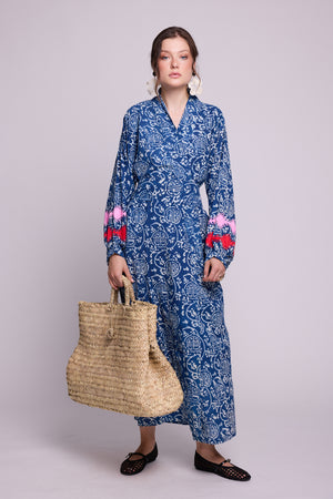 A woman modeling a long balloon sleeved indigo blue and white patterned handblock printed kimono by Kleed, 100% chemical free, with red and pink embroidered details on the cuffs, holding a large woven straw tote bag against a solid light gray background.
