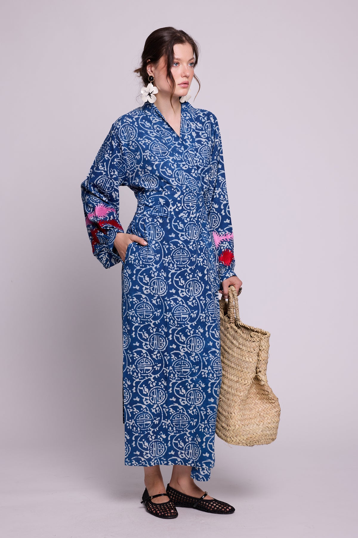 A woman wears KLEED’s 100% chemical-free, hand block-printed indigo blue and white maxi kimono with pink and red embroidered balloon sleeves, styled with a straw tote bag against a light grey background.