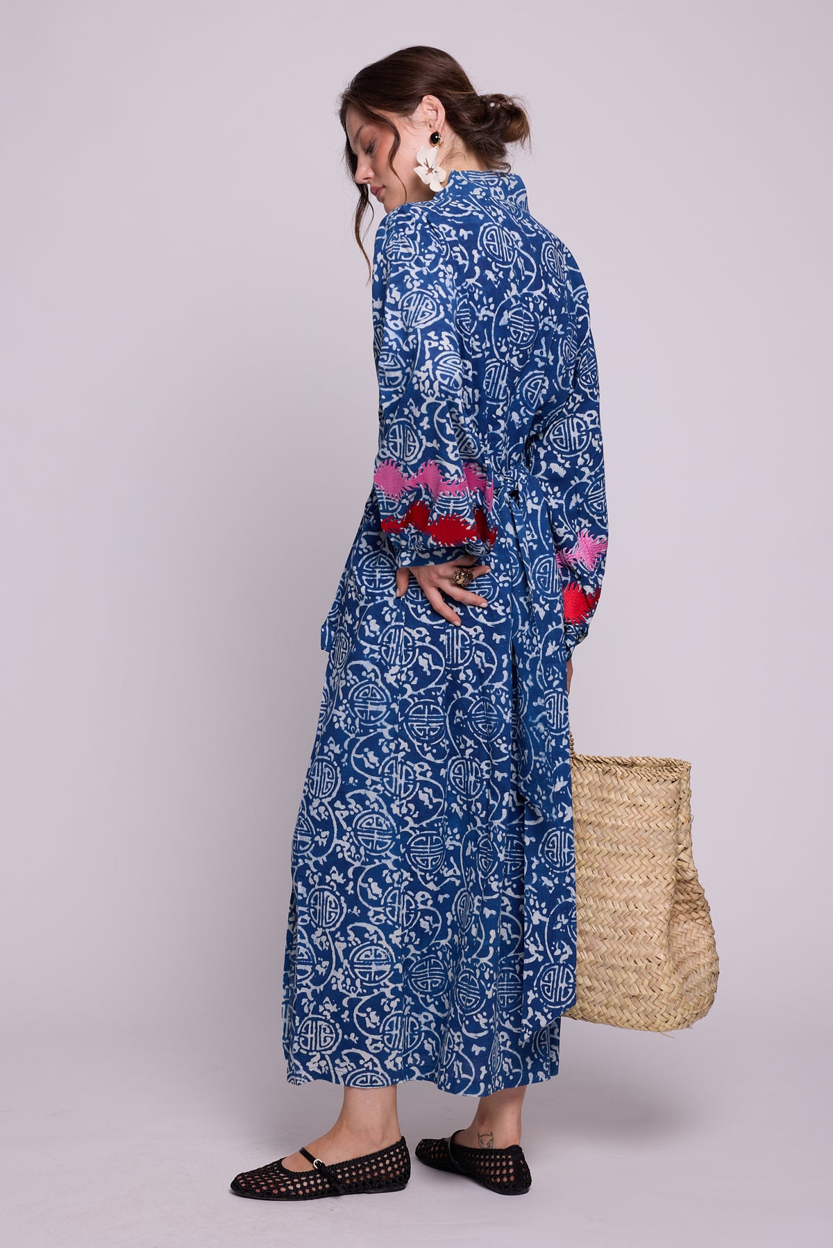 A woman models KLEED’s 100% chemical-free, hand block-printed indigo blue and white Indira Bloom Kimono from the back, featuring pink and red embroidered balloon sleeves and a matching belt, styled with a straw tote bag.