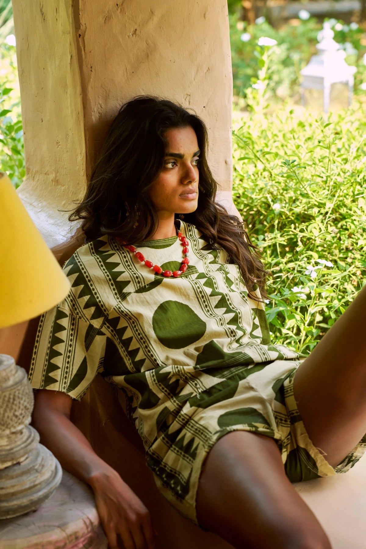 Kaner Khaki Geo Pyjama by KLEED, styled on a model reclining by a sunlit window in a green geometric cotton pyjama set with matching shorts and red beaded necklace.