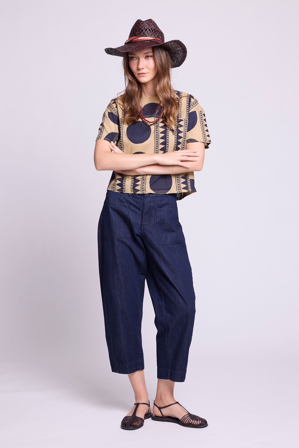 Studio photo of a model wearing the top of the Kaner Midnight Geo Pyjama by Kleed, in a beige base with midnight blue geometric motifs, styled with baggy denim trousers, a woven hat and black sandals.