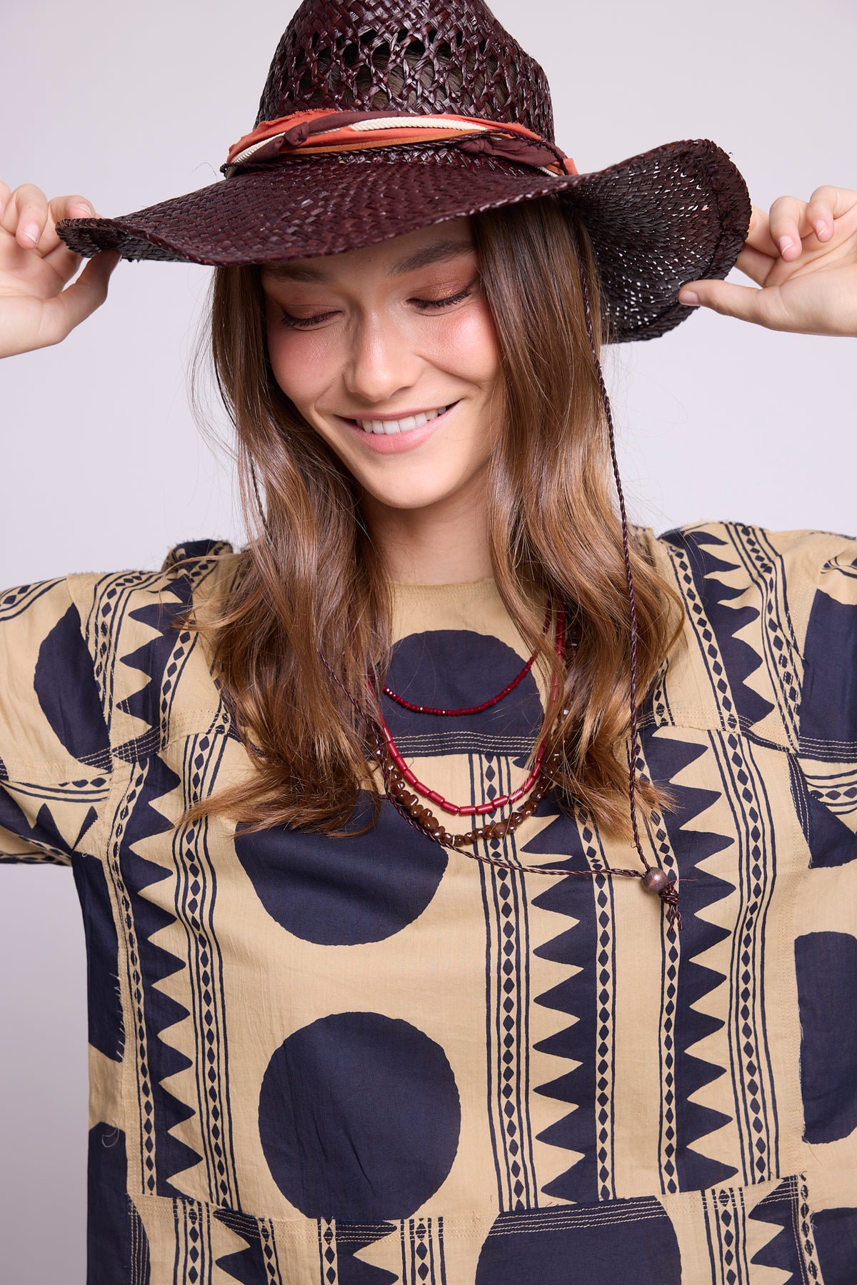 Close-up studio photo of a smiling model holding a woven hat, wearing the top of the Kaner Midnight Geo Pyjama by Kleed, showing the beige base and midnight blue geometric motifs in detail, styled with red beaded necklaces.