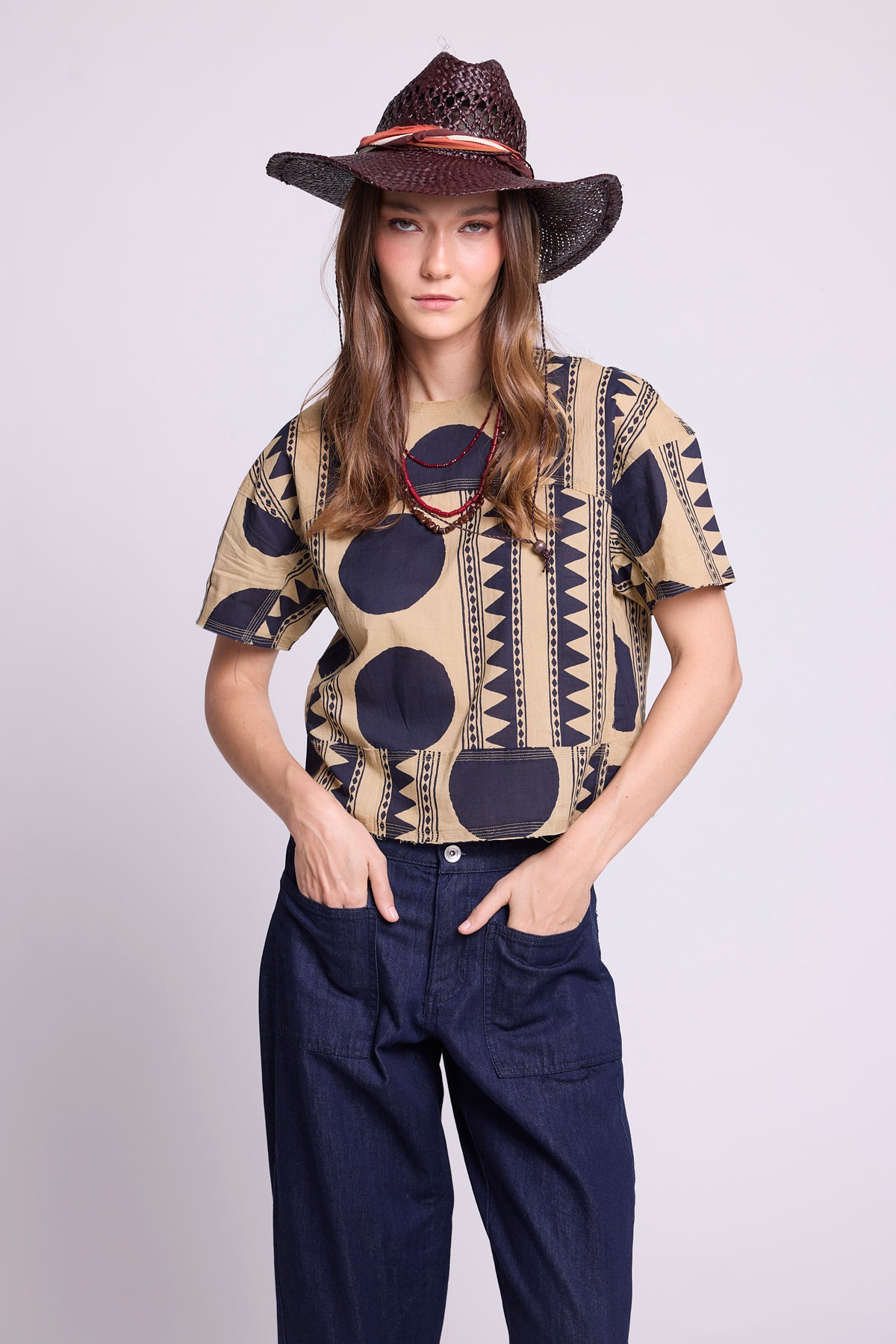 Close-up studio photo of a model wearing the top of the Kaner Midnight Geo Pyjama by Kleed, in a beige base with midnight blue geometric motifs, styled with baggy denim trousers, a woven hat and red beaded necklace, hands in pockets.