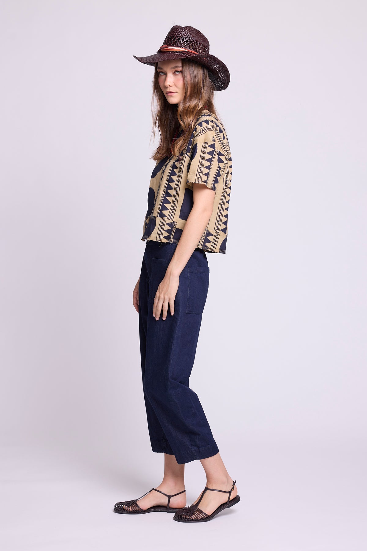 Side view studio photo of a model wearing the top of the Kaner Midnight Geo Pyjama by Kleed, in a beige base with midnight blue geometric motifs, styled with baggy denim trousers, a woven hat and black woven sandals.
