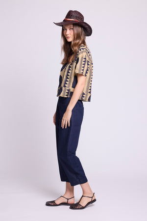 Side view studio photo of a model wearing the top of the Kaner Midnight Geo Pyjama by Kleed, in a beige base with midnight blue geometric motifs, styled with baggy denim trousers, a woven hat and black woven sandals.