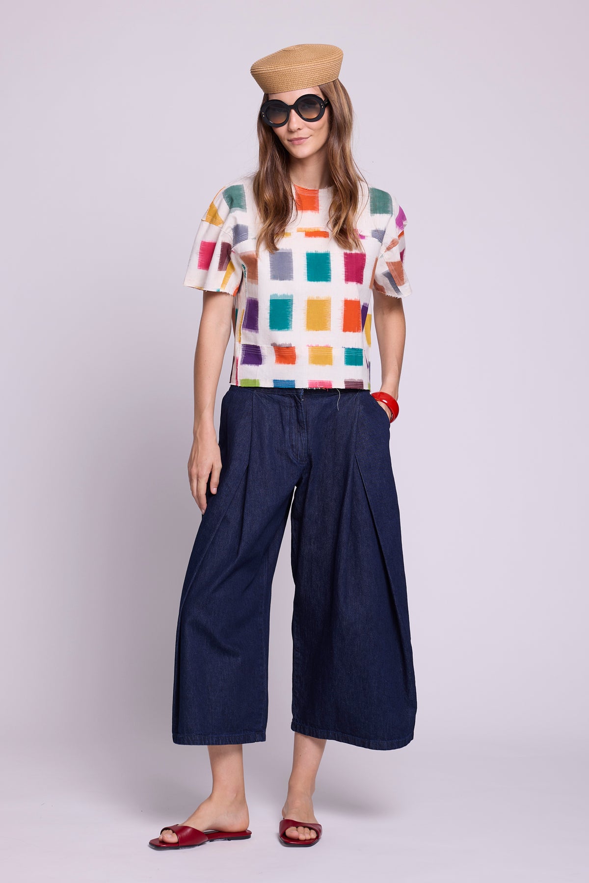 Studio photo of a model in a relaxed pose with one hand in her pocket, wearing the top of the Kaner Multisquares Pyjama by Kleed, in hand-woven cotton ikat with colourful squares, paired with dark denim culottes, a straw hat, oversized sunglasses and red leather sandals.