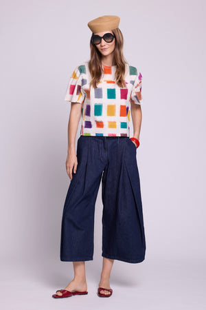 Studio photo of a model in a relaxed pose with one hand in her pocket, wearing the top of the Kaner Multisquares Pyjama by Kleed, in hand-woven cotton ikat with colourful squares, paired with dark denim culottes, a straw hat, oversized sunglasses and red leather sandals.