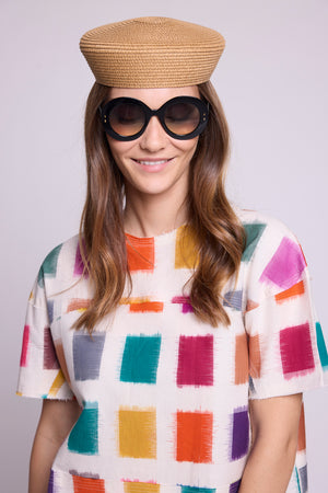 Close-up studio photo of a smiling model wearing the top of the Kaner Multisquares Pyjama by Kleed, showing the multicoloured ikat squares on a white cotton base in detail, styled with a straw hat and round black sunglasses.