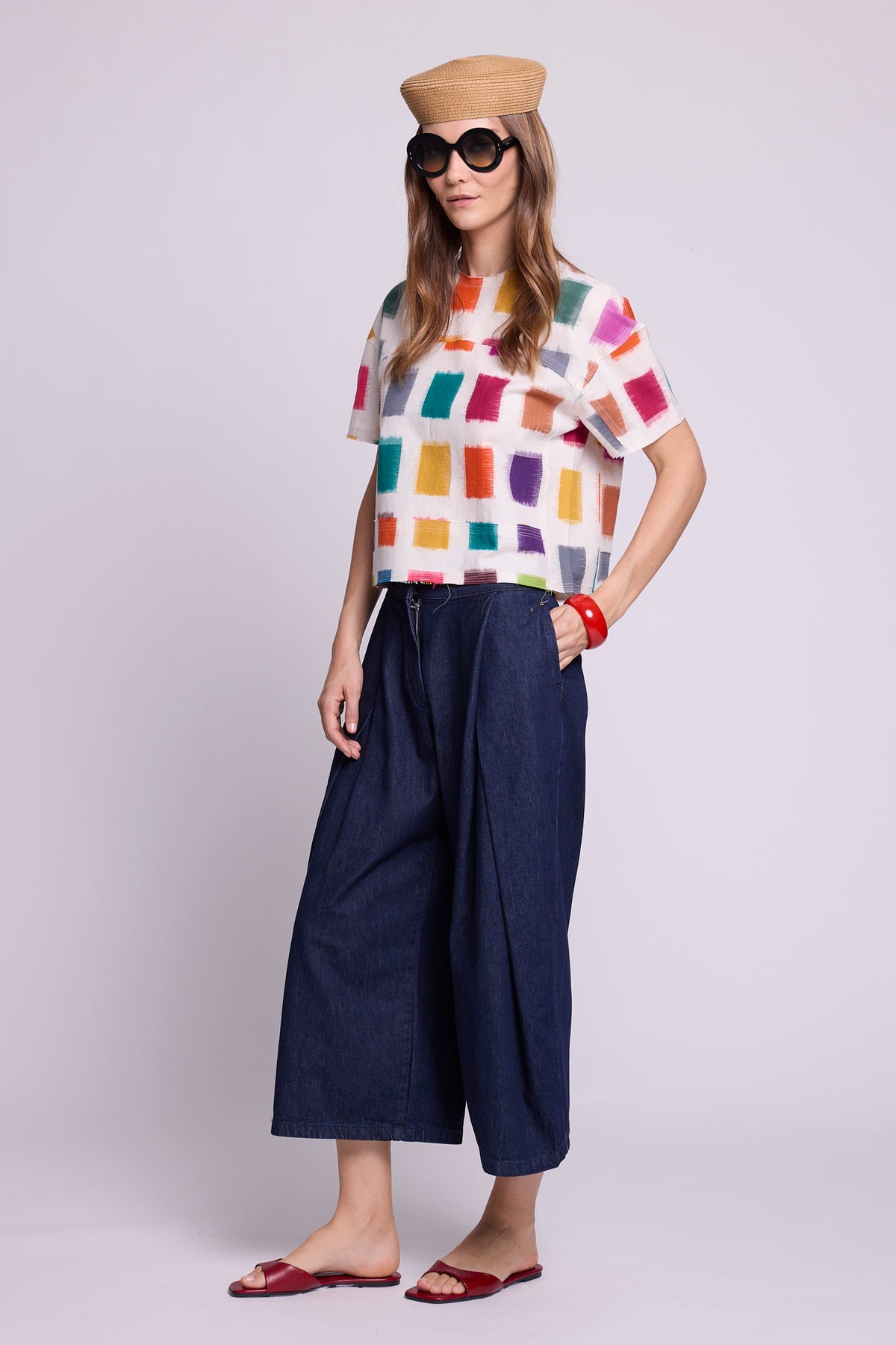 Three-quarter view studio photo of a model wearing the top of the Kaner Multisquares Pyjama by Kleed, in multicoloured cotton ikat squares on a white base, styled with wide-leg cropped denim trousers, a straw hat, sunglasses, a red bangle and red sandals.