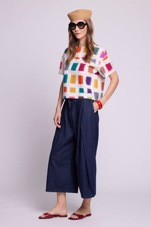 Three-quarter view studio photo of a model wearing the top of the Kaner Multisquares Pyjama by Kleed, in multicoloured cotton ikat squares on a white base, styled with wide-leg cropped denim trousers, a straw hat, sunglasses, a red bangle and red sandals.