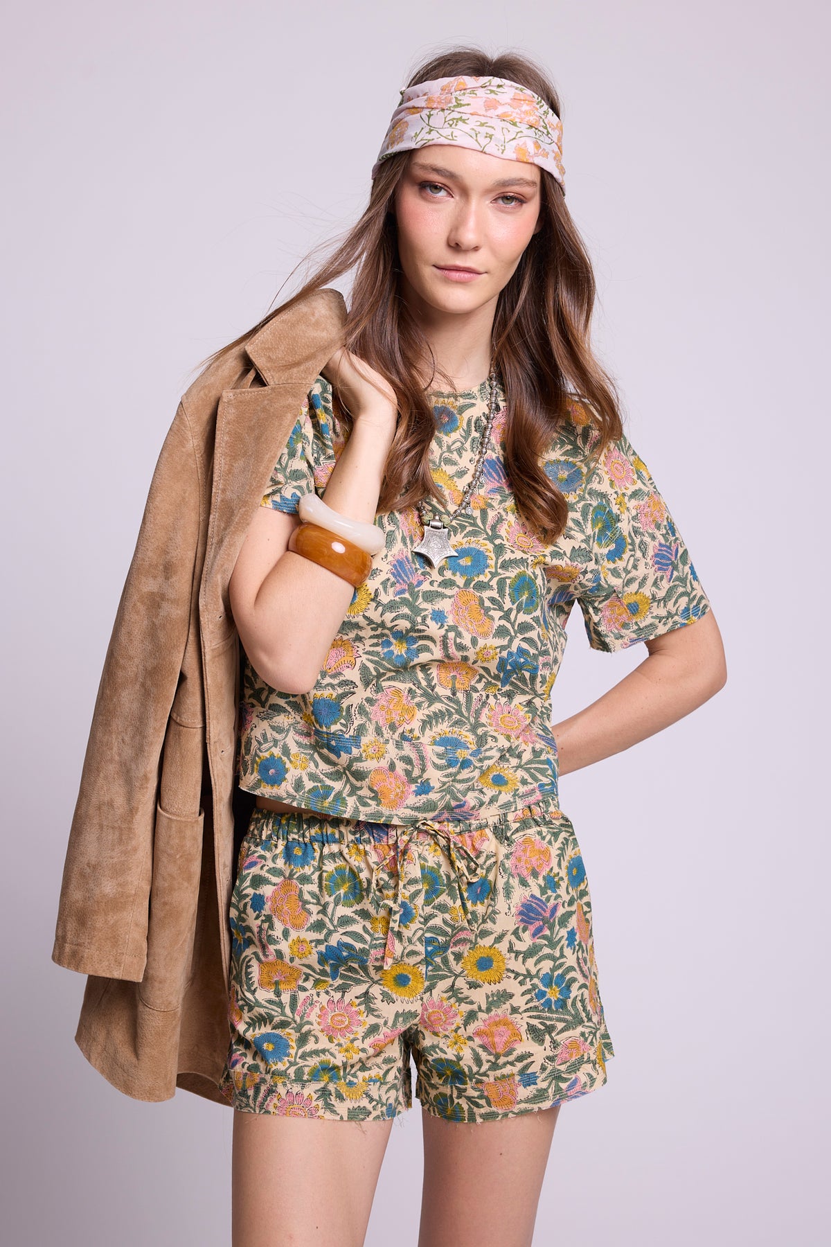 Studio photo of a model with a camel suede jacket slung over her shoulder, wearing the Kaner Palash Pyjama by Kleed, a top and drawstring shorts set in cream cotton with blue, pink and yellow floral print, styled with a floral headscarf and chunky bangles.