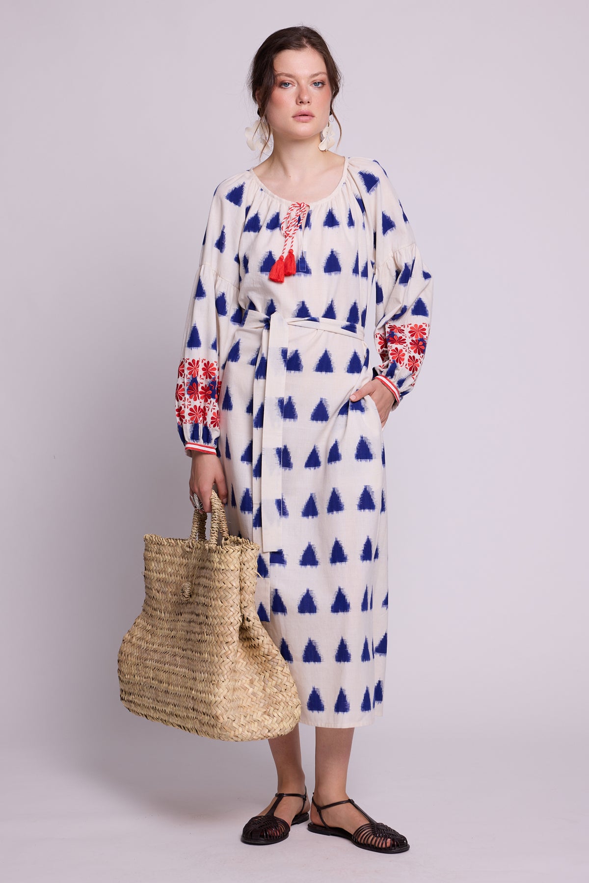 Model wearing the KLEED Saint Tropez Kaftan in cotton linen with blue geometric motifs and red and blue embroidered sleeves, styled with statement earrings, black flat sandals and a large woven straw basket.