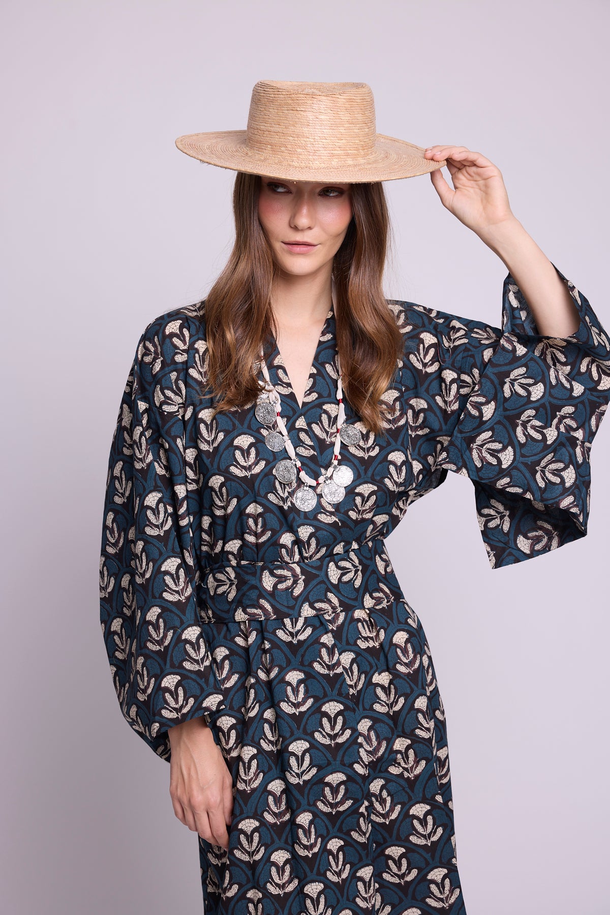 close up of model wearing krisant naya kimono by Kleed.. 100% hand blockprinted, flower motifs and white black and blue. Holding a straw hat, and wearinga silver boho necklace.