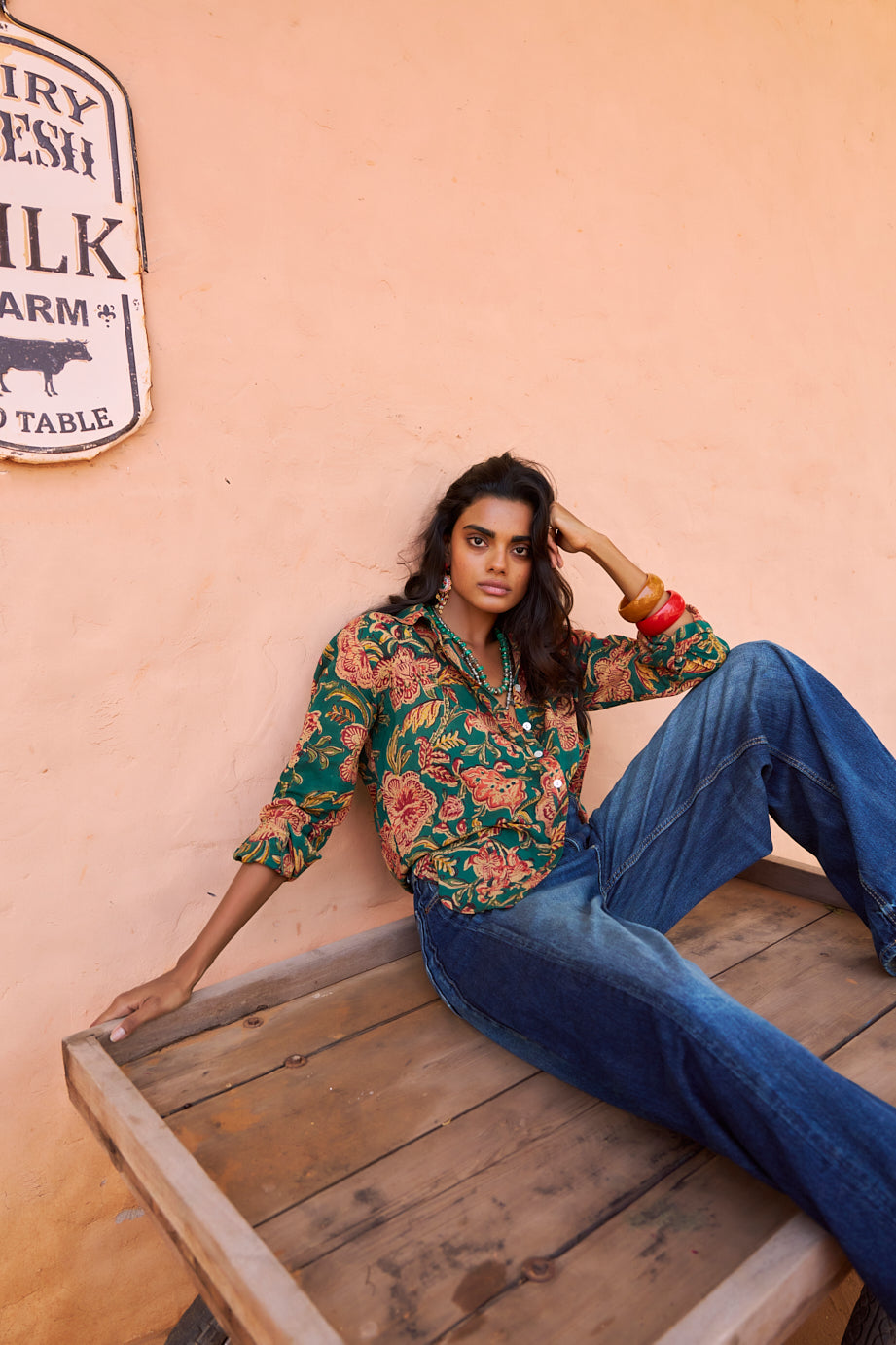 Model seated on a rustic wooden platform against a peach-toned wall, wearing the KLEED Lotus Green Shirt in 100% hand block printed cotton with mother-of-pearl buttons, styled with relaxed blue denim.