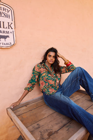 Model seated on a rustic wooden platform against a peach-toned wall, wearing the KLEED Lotus Green Shirt in 100% hand block printed cotton with mother-of-pearl buttons, styled with relaxed blue denim.