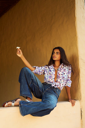 Model wearing the KLEED Lotus White Shirt in 100% cotton, hand block printed with lilac, blue, turquoise and burgundy floral motifs, styled tied at the waist with relaxed blue jeans.