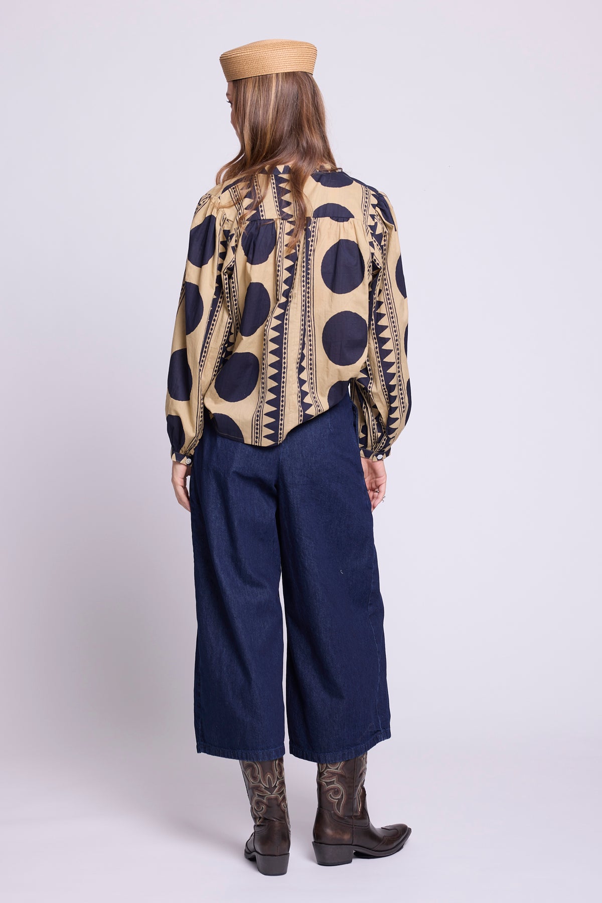 A model shown from the back wearing a beige and navy patterned shirt, the midnight geo balloon shirt by kleed, with dark blue wide-leg cropped pants, brown cowboy boots, and a tan beret, set against a plain light gray studio background.