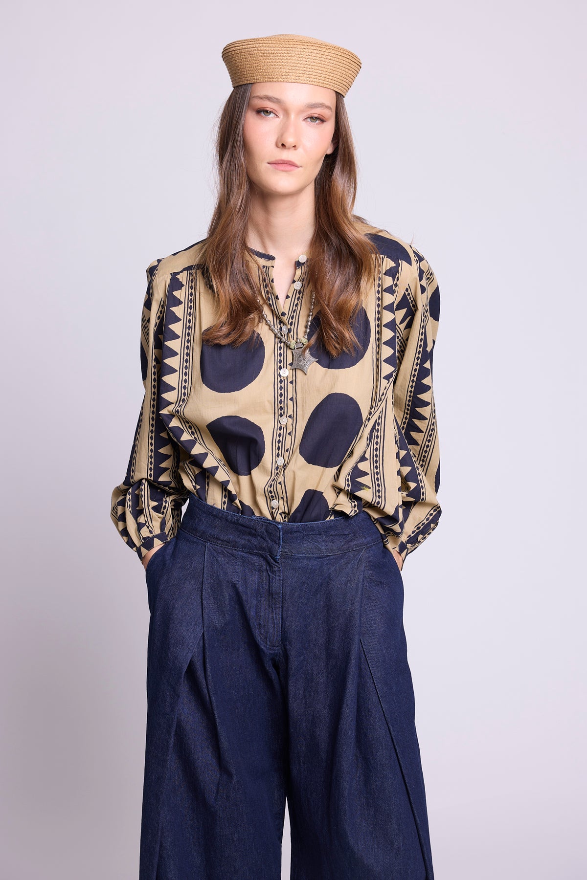 A woman modeling a beige and navy patterned shirt, the midnight geo balloon shirt by kleed, with geometric prints, paired with dark blue wide-leg pants and a beige straw hat, against a light gray studio background.