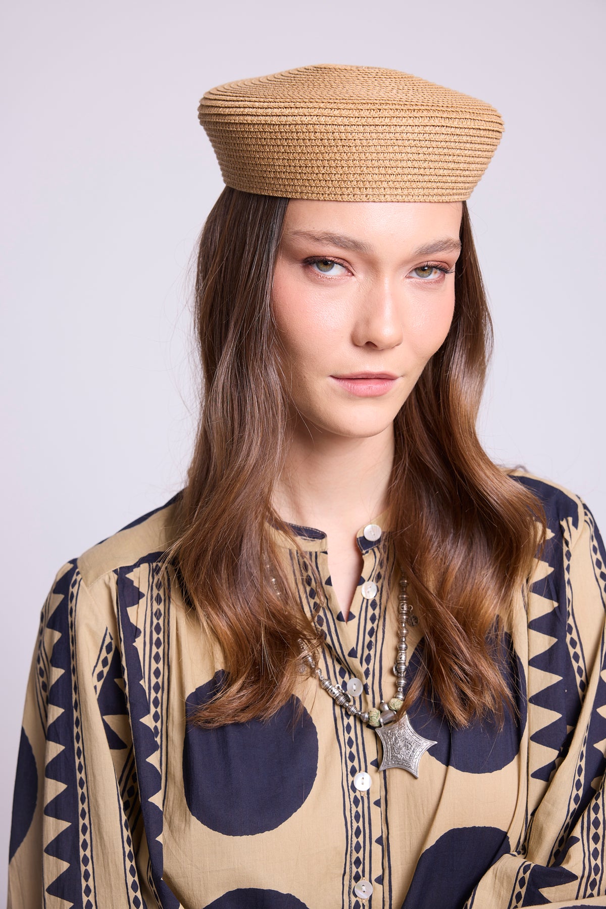 A woman wearing a woven tan beret-style hat and a beige patternedkleed shirt the midnight geo balloon shirt, in 100% cotton, with dark navy geometric motifs, photographed against a light gray studio background.