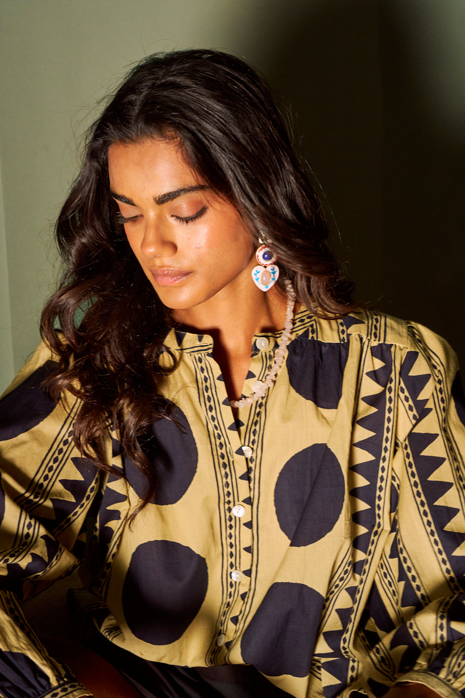 Close-up of a model wearing the KLEED Midnight Geo Balloon Shirt in 100% cotton, featuring bold black geometric and circular motifs on a beige background, styled with colourful statement earrings and softly waved dark hair.